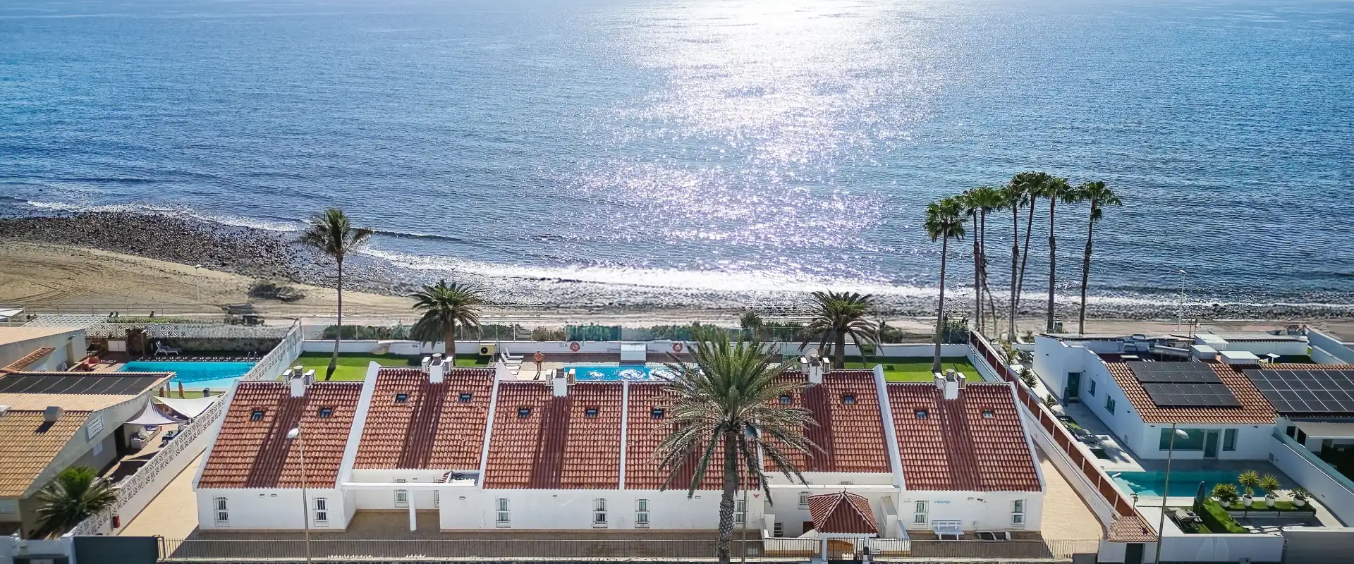 Viviendas con techos rojos frente al oc&eacute;ano, rodeadas de palmeras y piscinas.