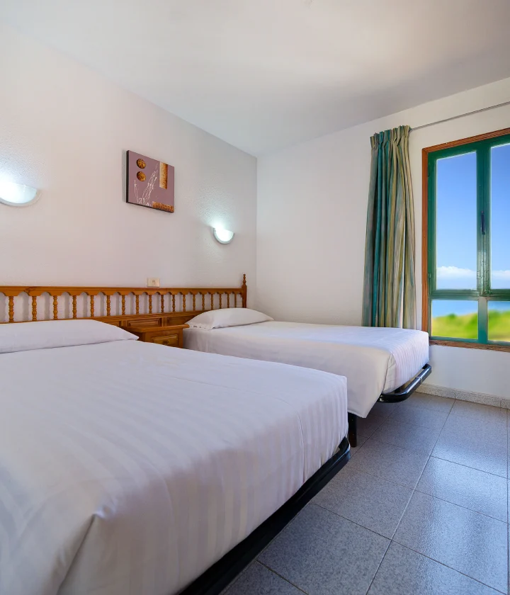 Habitaci&oacute;n con dos camas individuales, l&aacute;mparas de pared y ventana con vista al exterior.