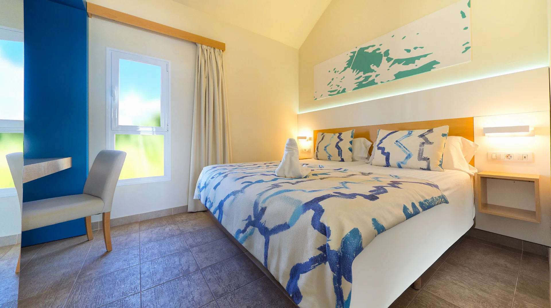 Habitaci&oacute;n luminosa con cama doble, decoraci&oacute;n azul, ventana y mesa con silla.