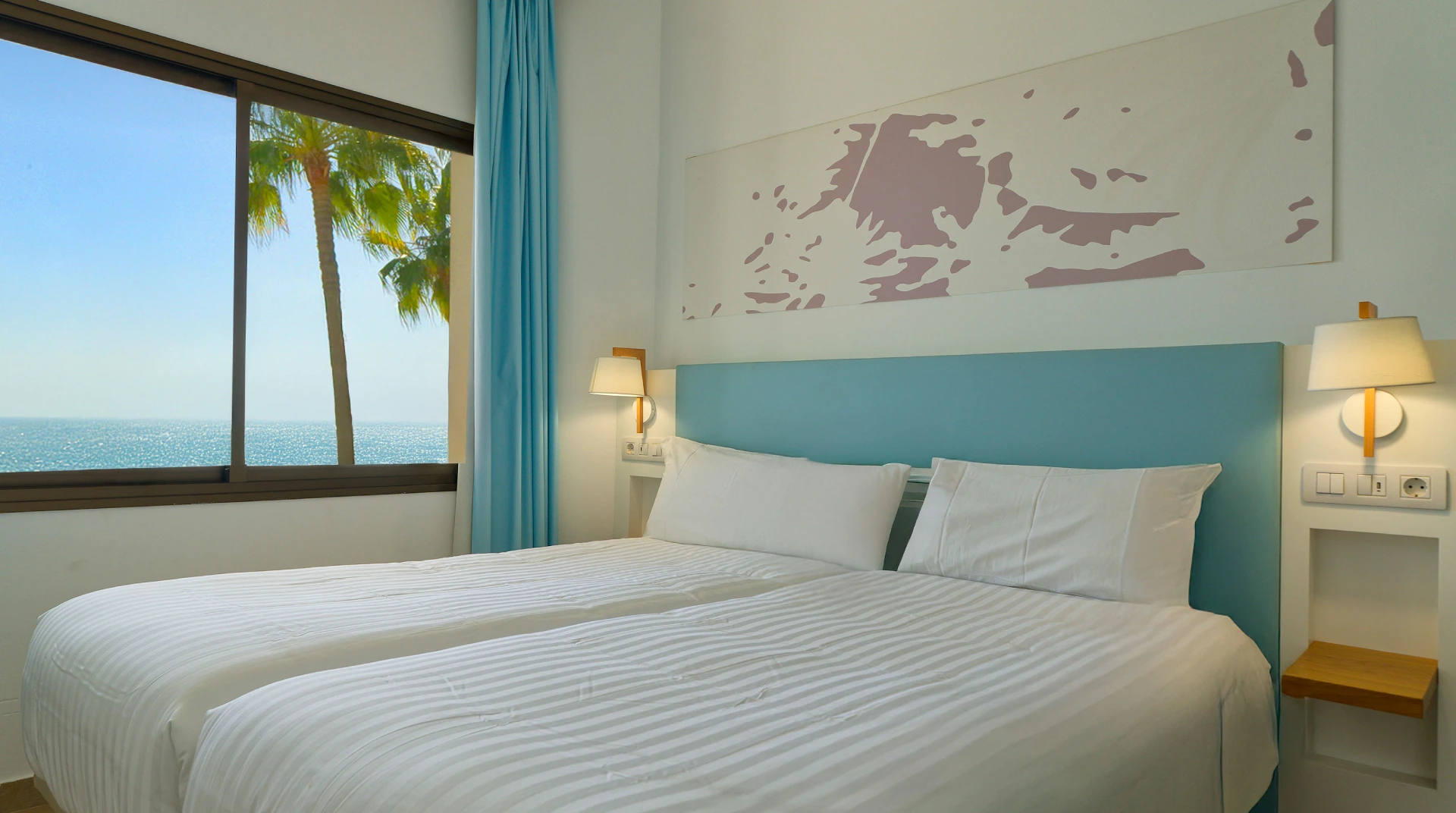 Habitaci&oacute;n luminosa con camas gemelas, decoraci&oacute;n azul, ventana con vista al mar y palmeras.