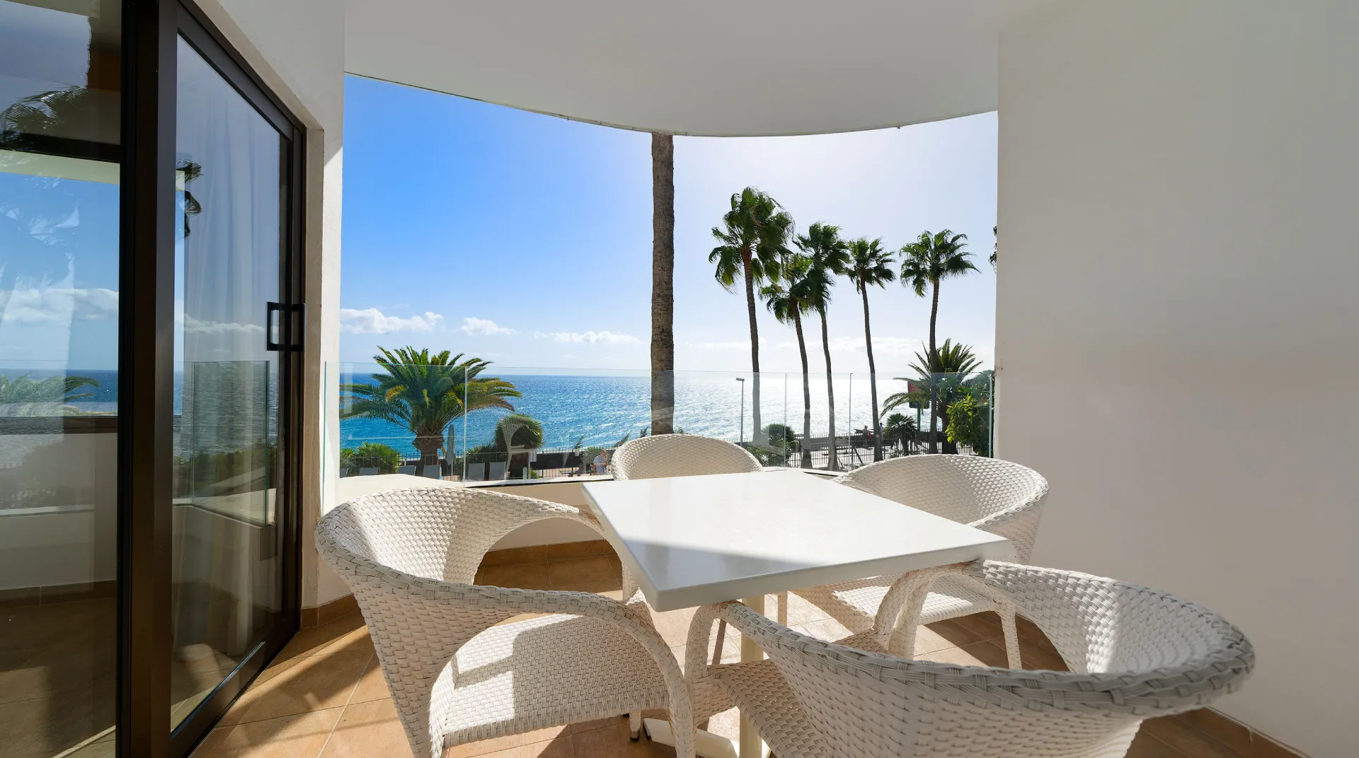 Balc&oacute;n con muebles blancos frente al mar, palmeras y cielo despejado.