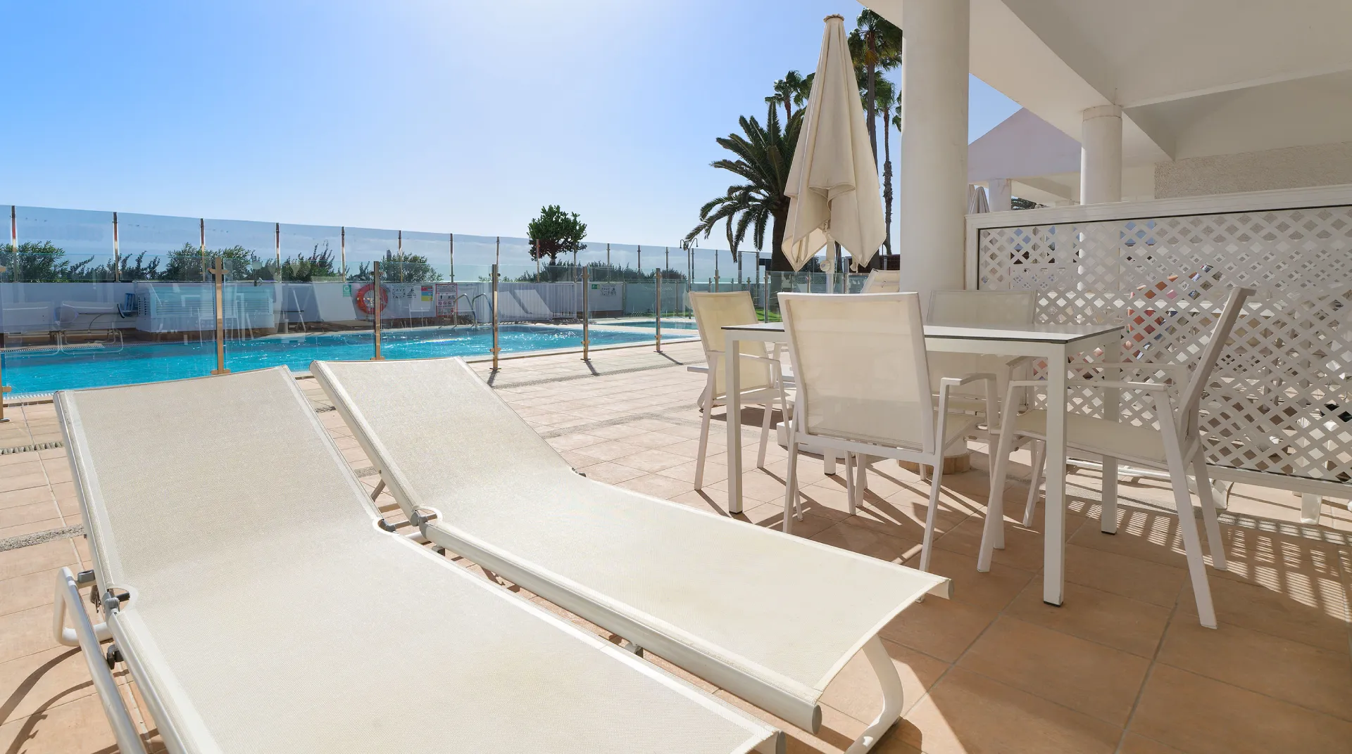 Terraza con tumbonas, mesa, sillas y piscina al fondo, rodeada de una valla de vidrio.