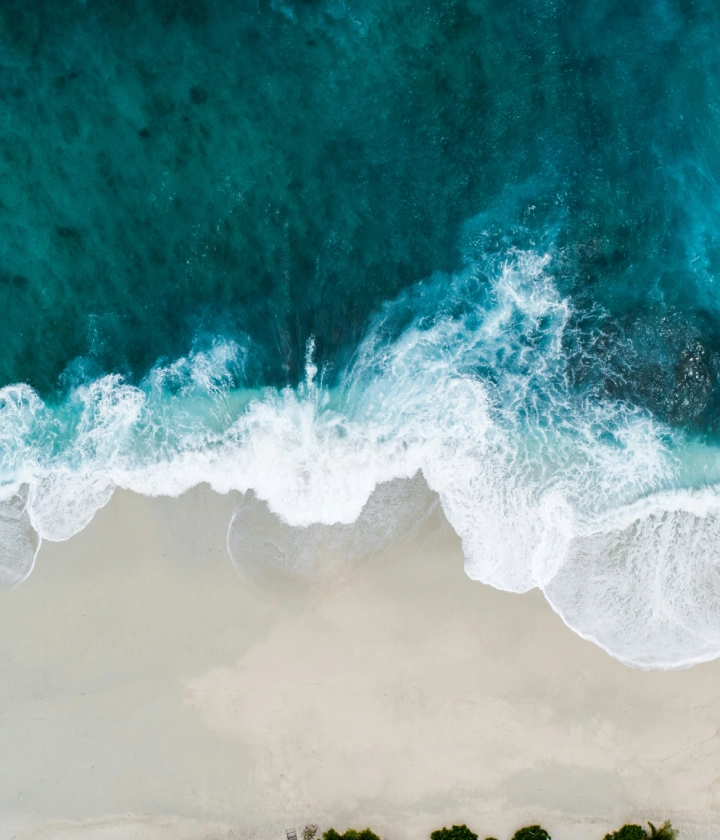 Playa vista aérea: olas azules rompen sobre arena blanca; vegetación al fondo.
