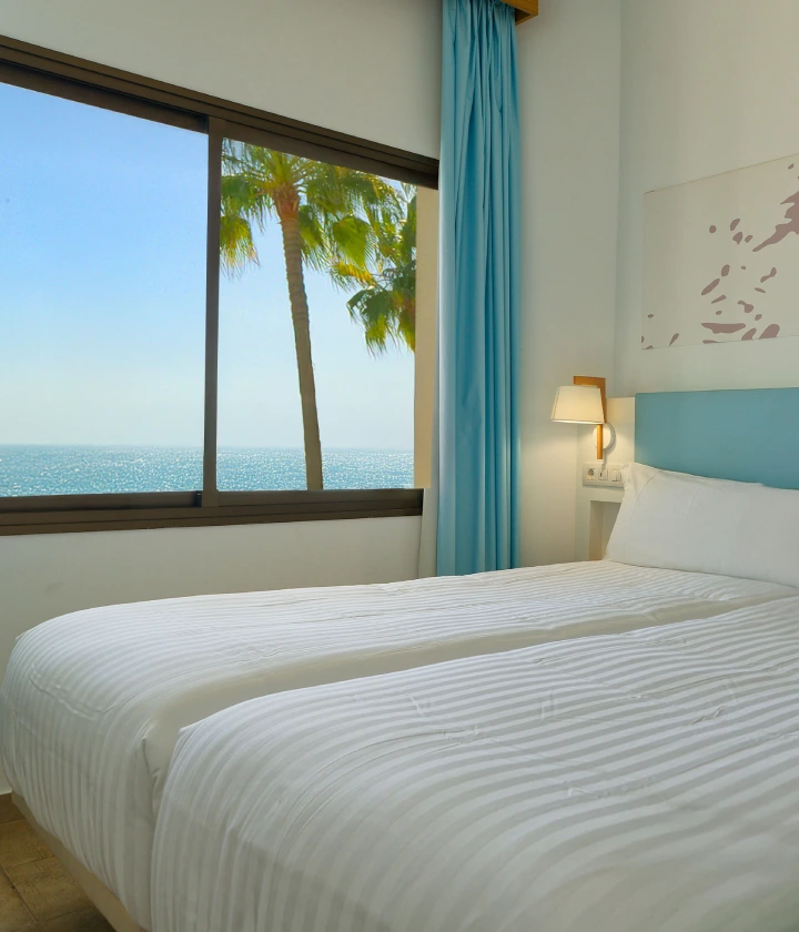 Habitaci&oacute;n con dos camas, ventana mostrando vista al mar y una palmera. Decoraci&oacute;n en tonos azules.