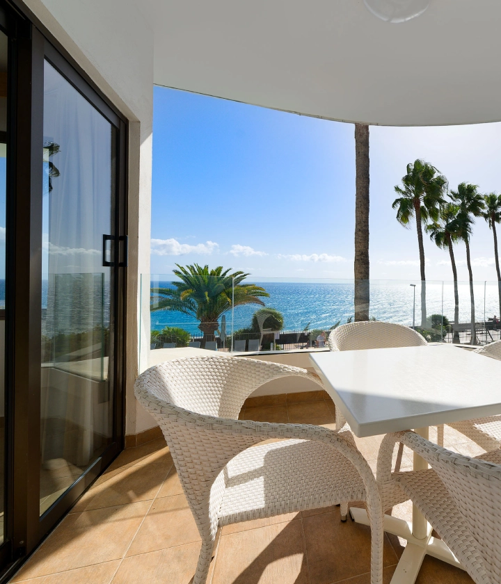 Balc&oacute;n con mesa y sillas de mimbre, vista al mar, palmeras y cielo azul.