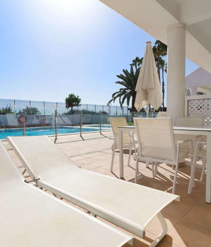 Terraza con tumbonas, mesa y sillas blancas cerca de una piscina, rodeada de palmeras.