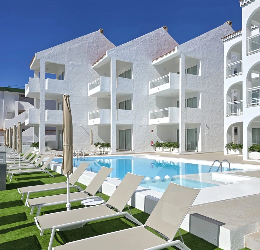 Edificio blanco con balcones y piscina; tumbonas alineadas y cielo despejado.