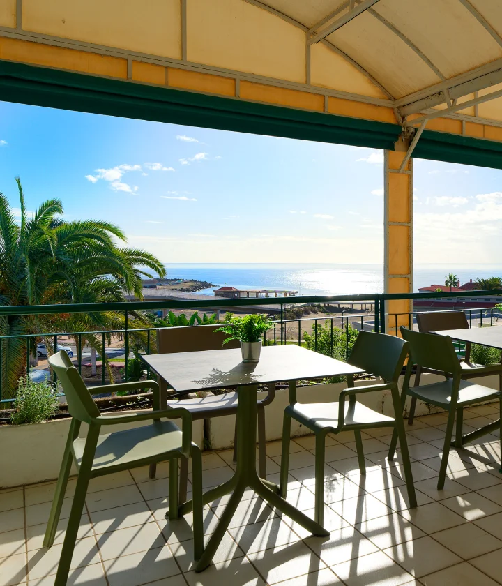 Terraza con mesa y sillas, vistas al mar, palmeras y cielo azul.