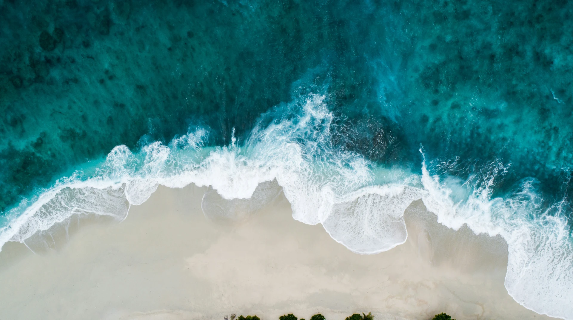 Vista a&eacute;rea de olas rompiendo en una playa de arena clara y agua turquesa.