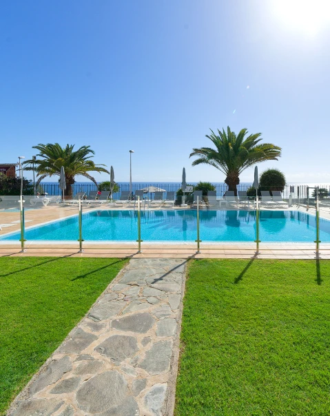 Piscina rodeada de césped, con palmeras y vista al mar bajo un cielo despejado.