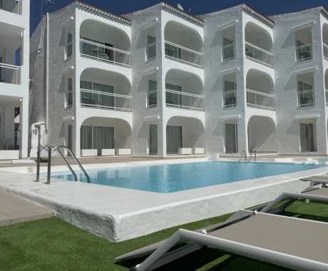 Edificio blanco con piscina y tumbonas en primer plano.