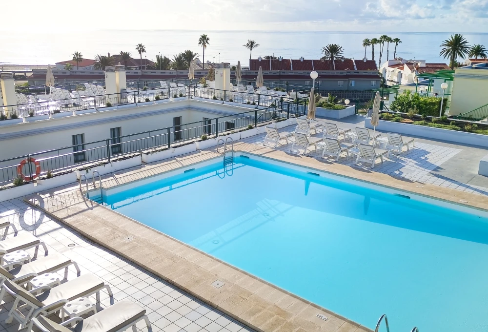 Piscina en una terraza con tumbonas y sombrillas, vista al mar y palmeras al fondo.