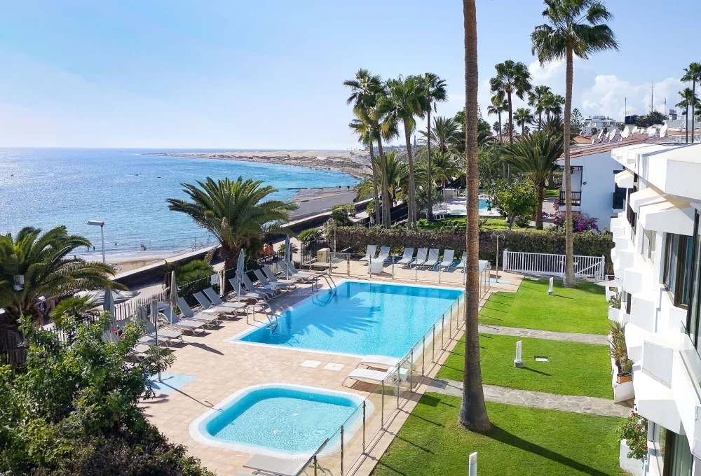 Piscina con vistas al mar y jardines, rodeada de palmeras y tumbonas.