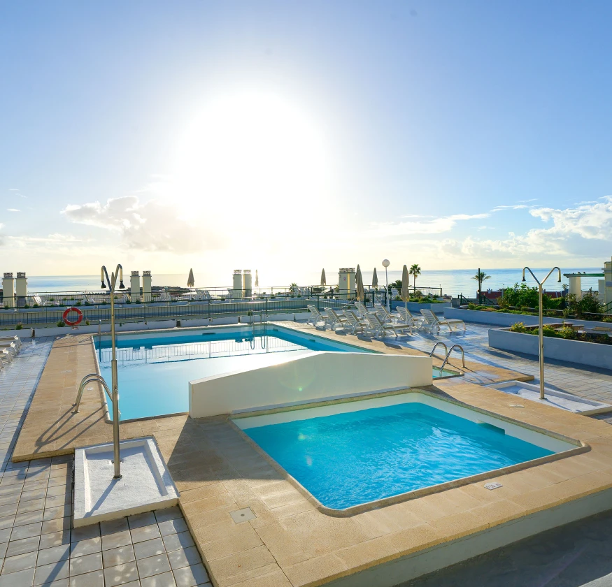 Piscina al aire libre con tumbonas bajo un cielo soleado y vista al mar al fondo.