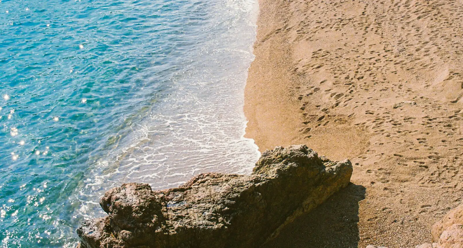 Playa con agua azul y rocas, arena con huellas, olas suaves en la orilla.