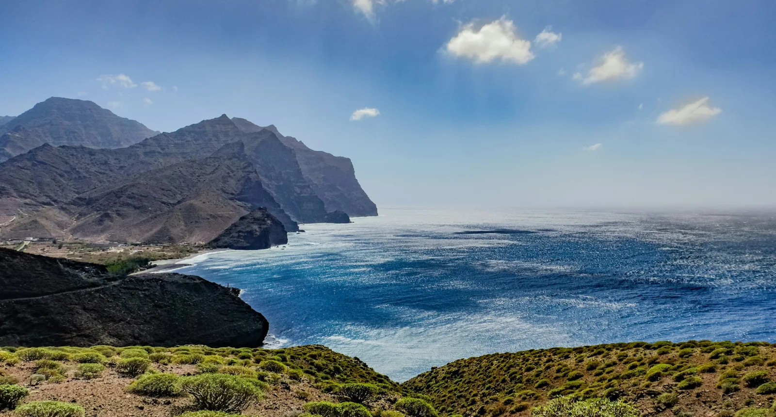 Costa rocosa con monta&ntilde;as al fondo, mar azul y cielo despejado con pocas nubes.