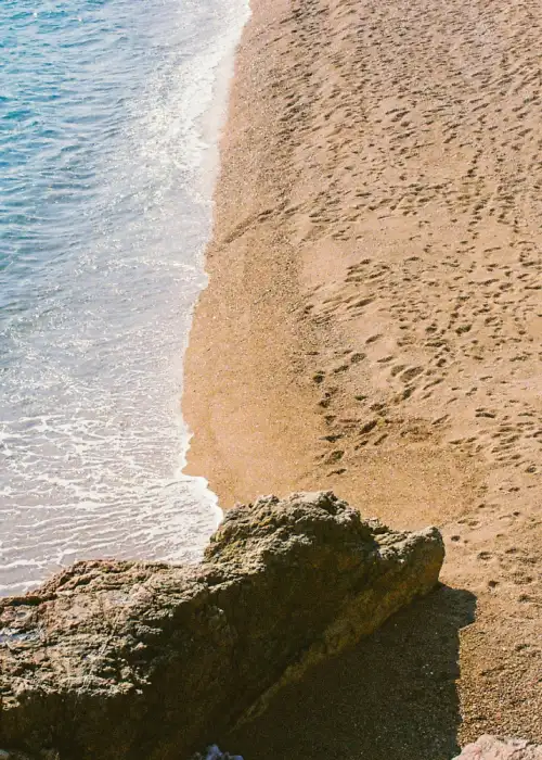 Playa con olas suaves, arena dorada y rocas, mostrando huellas a lo largo de la orilla.