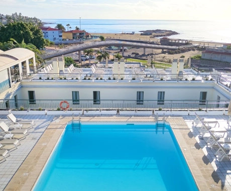 Piscina en una terraza con vista al mar y edificios, rodeada de sillas y vegetación.