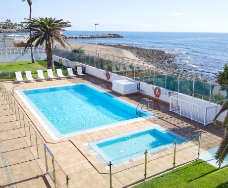 Piscina junto al mar con tumbonas, césped y vista a la playa rocosa y al horizonte azul.