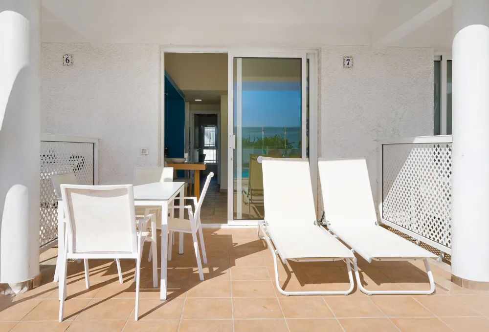 Terraza con dos tumbonas y mesa blanca, vista al mar a trav&eacute;s de la puerta corredera de vidrio.