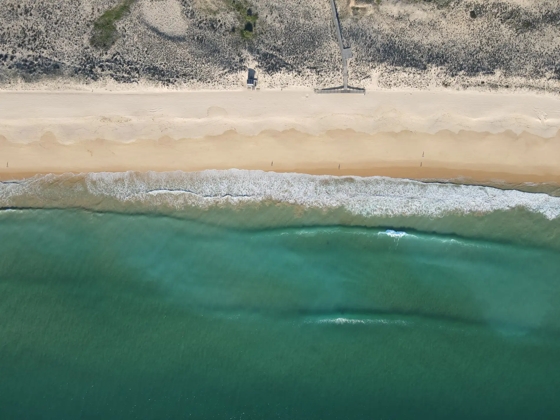 Aerial view of a beach with waves, sandy shore, and dunes in the background.