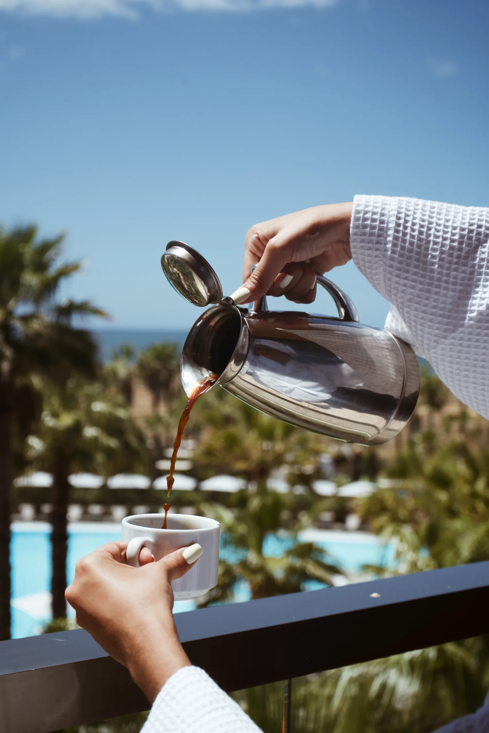 Pouring coffee from a pot into a cup with a scenic view of palm trees and a pool.