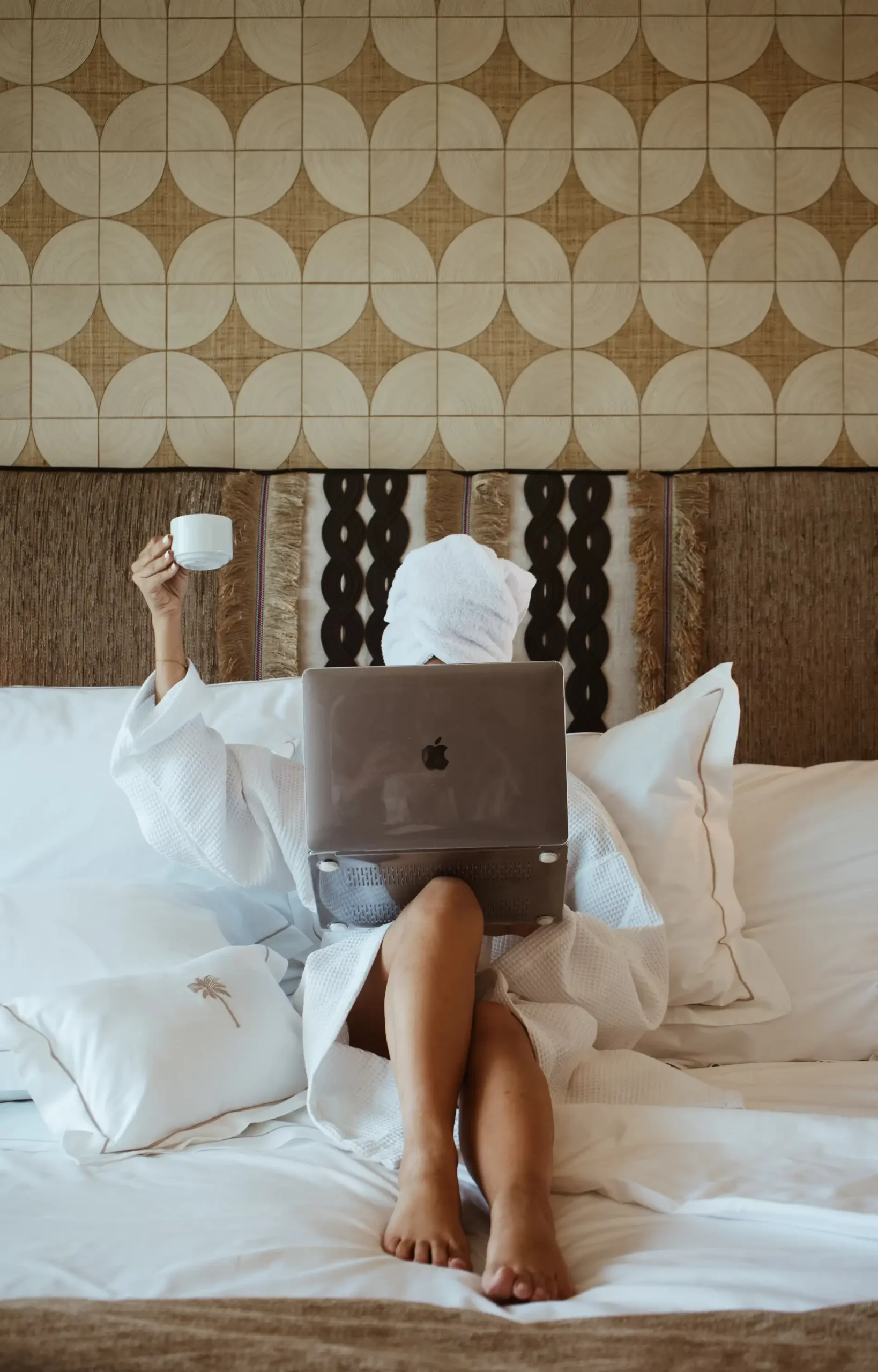 Person in robe on bed with laptop, holding a cup, wrapped in towel, decorative wall background.