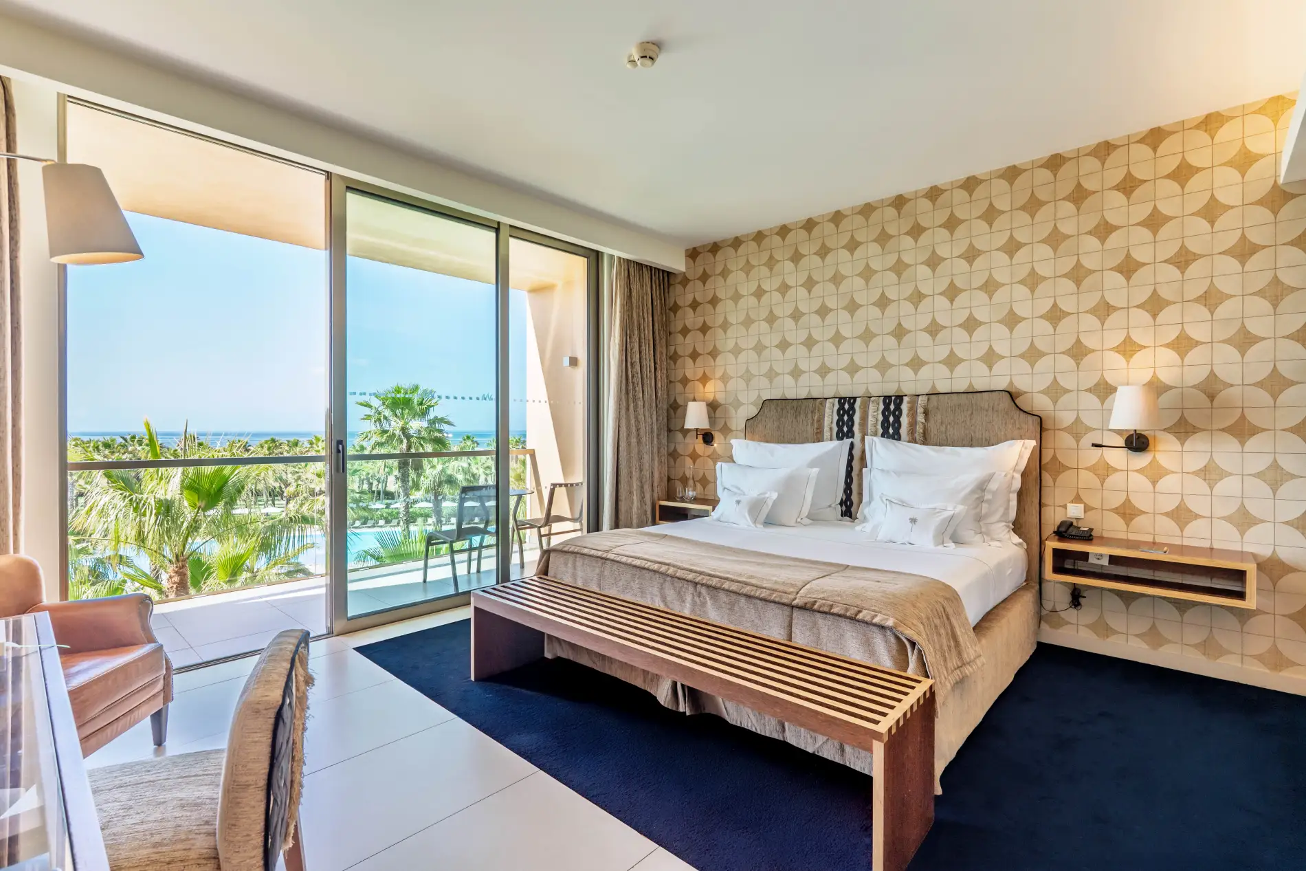 A modern hotel room with a bed, patterned wall, and balcony view of palm trees and the sea.