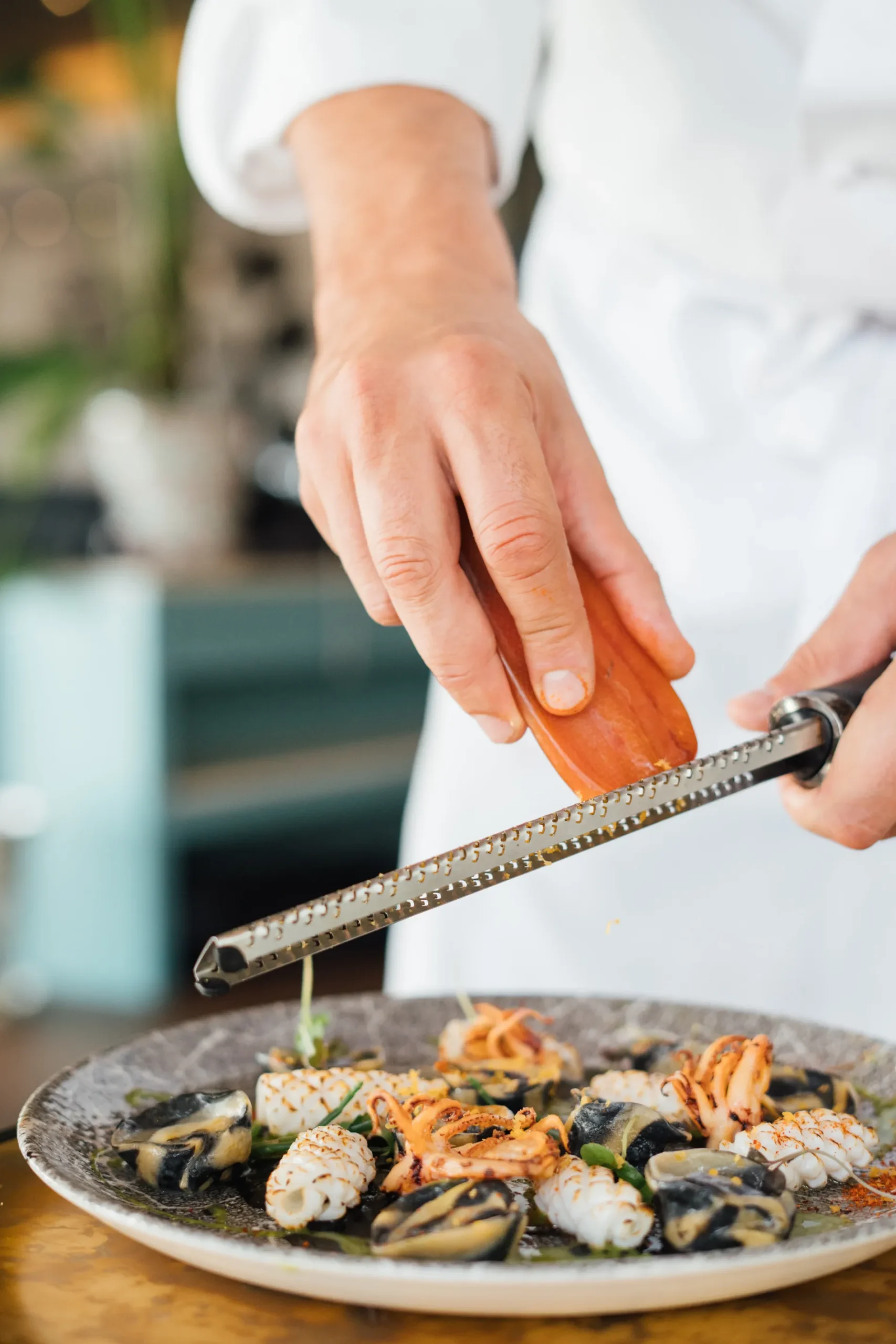 Chef grates over a plate of seafood and pasta in an artistic presentation.