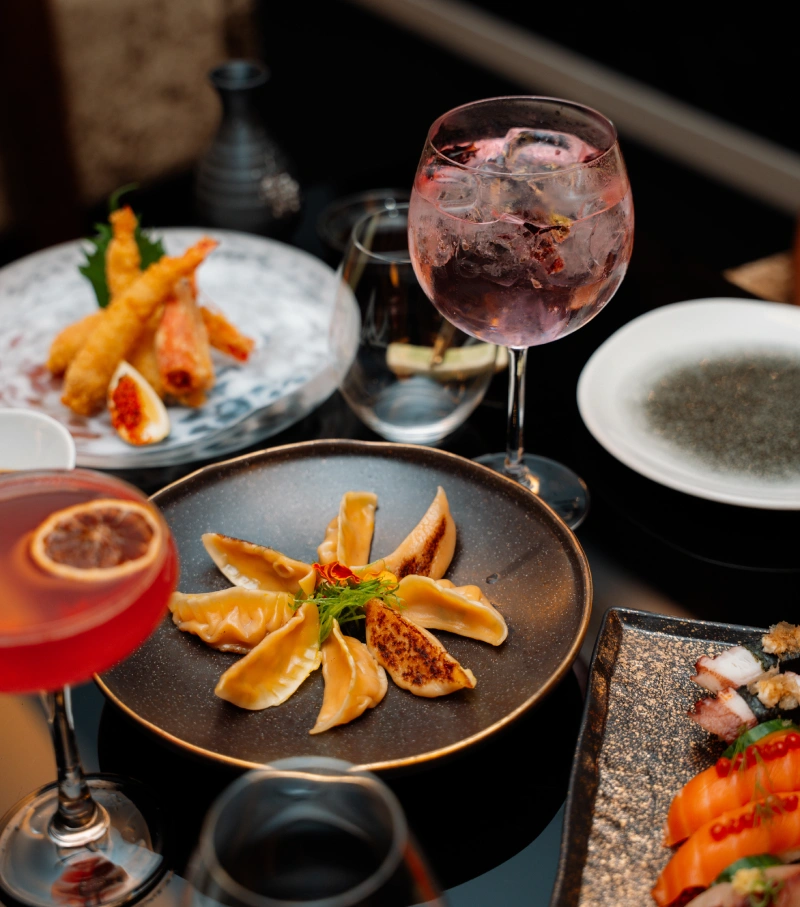 Dumplings and tempura with cocktails on a dark table setting.