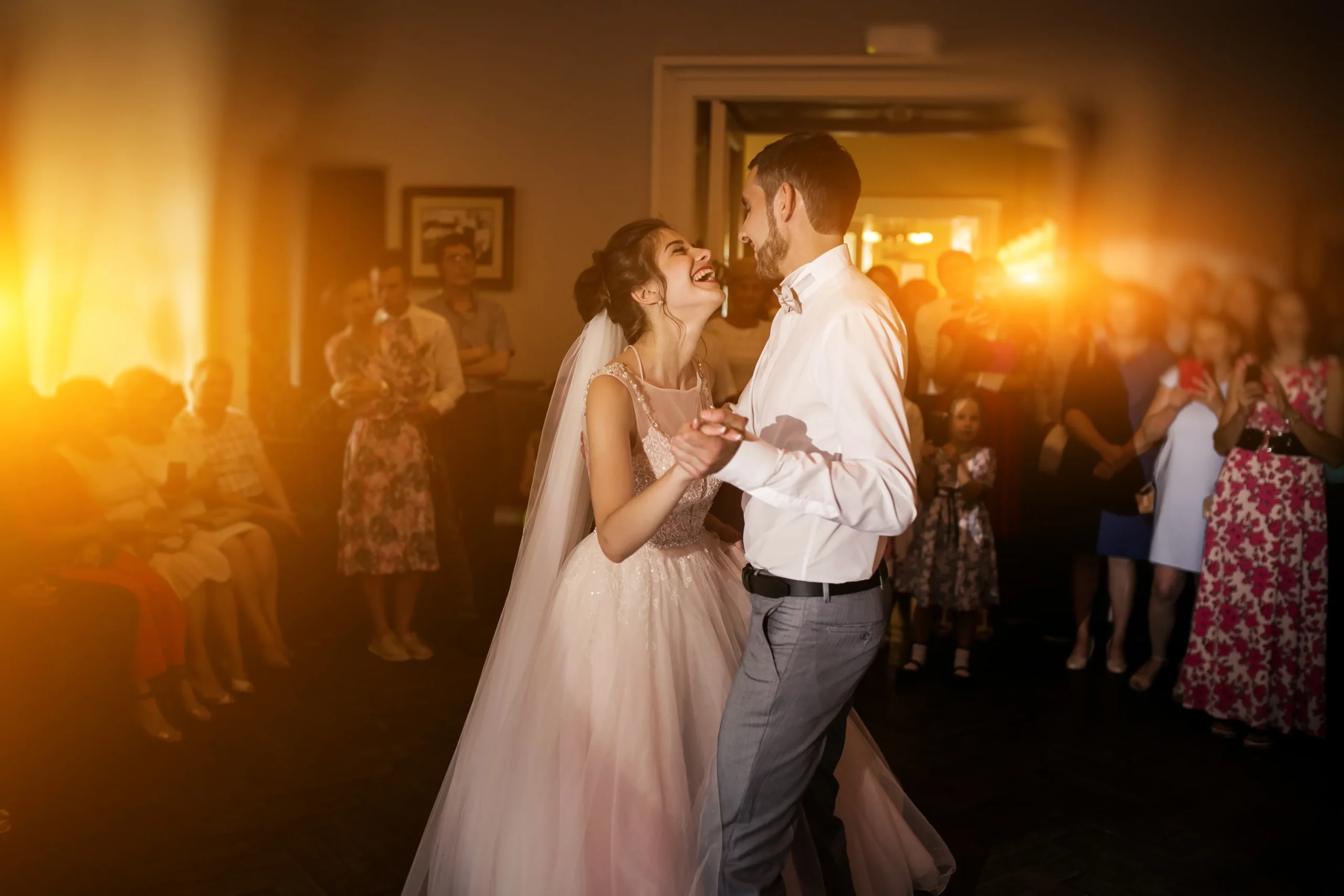 Pareja de reci&eacute;n casados bailando, rodeados de invitados y luces c&aacute;lidas.