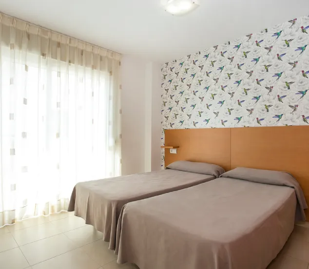 Habitación luminosa con dos camas individuales, papel tapiz de colibríes y cortinas claras.