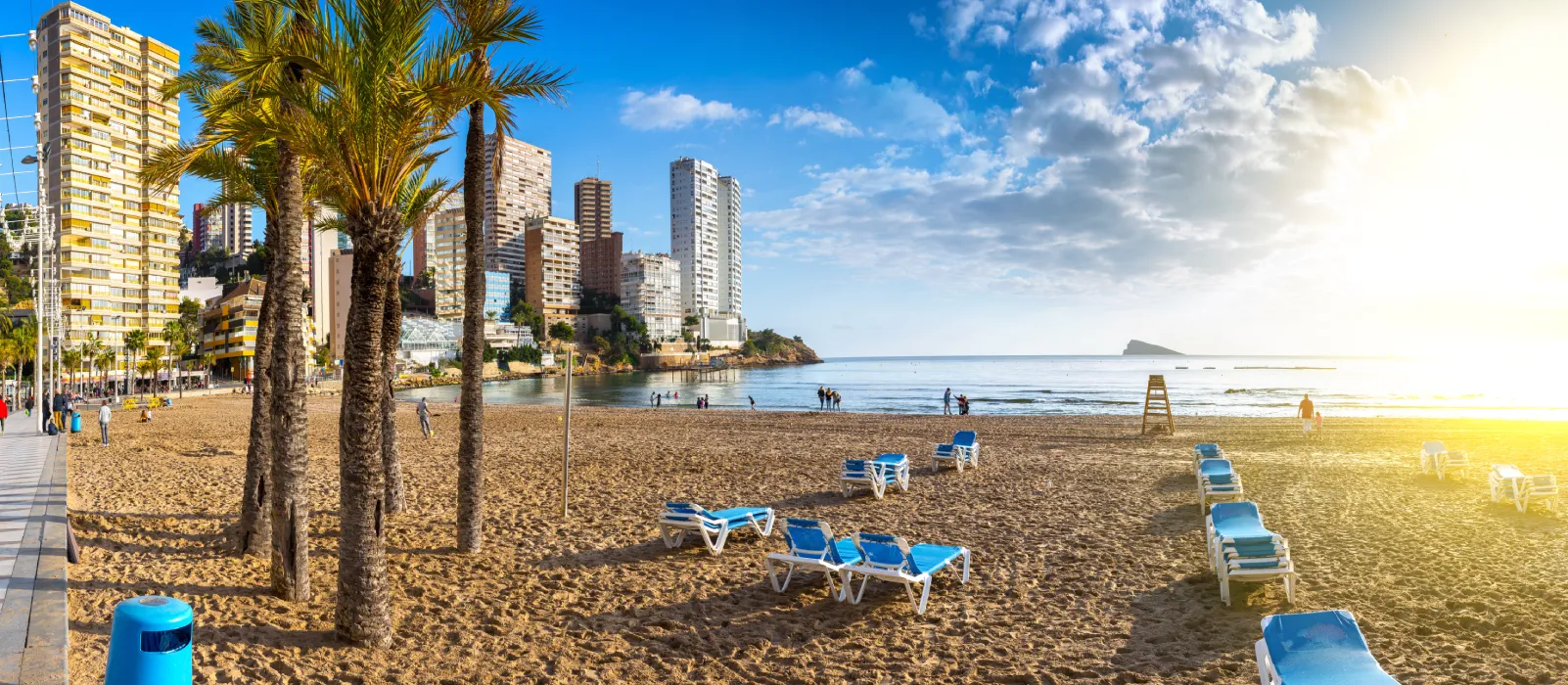 Playa con sombrillas y edificios altos al fondo bajo un cielo azul con algunas nubes.
