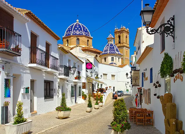 Calle pintoresca de un pueblo mediterráneo con casas blancas y una iglesia de cúpulas azules.