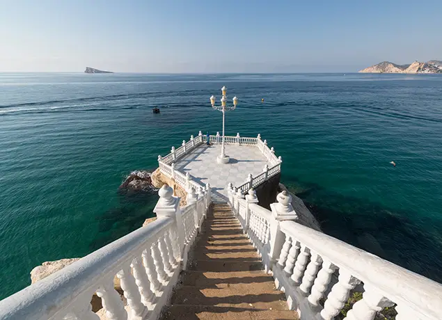 Escaleras blancas llevan a un mirador sobre el mar con farola y barandillas, cielo despejado.