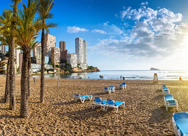 Playa con palmeras, tumbonas azules y edificios al fondo, cielo despejado al amanecer.