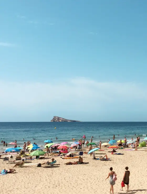 Playa concurrida con sombrillas coloridas, personas tomando sol y una isla al fondo.
