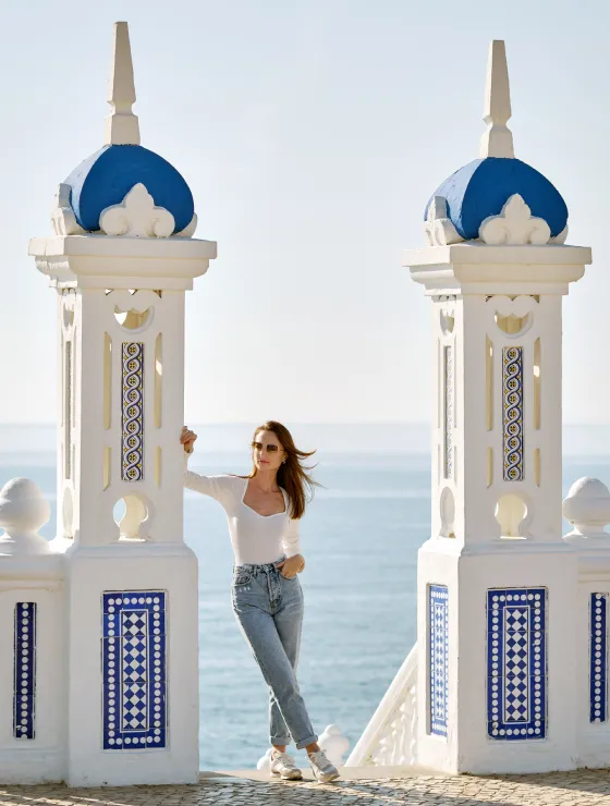 Mujer entre pilares blancos y azules frente al mar en un día soleado.