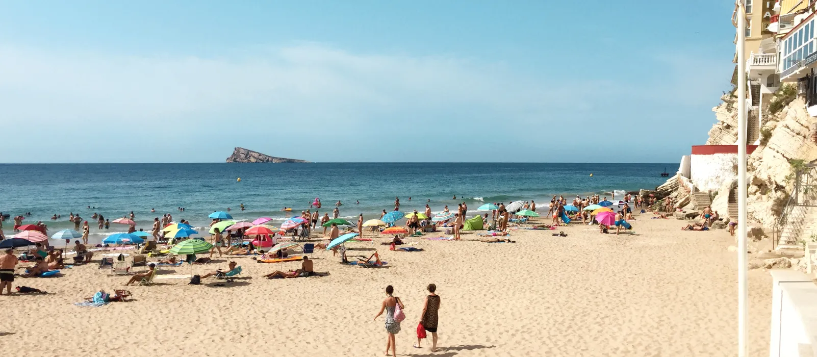 Playa concurrida con sombrillas de colores, gente nadando y una isla al fondo.