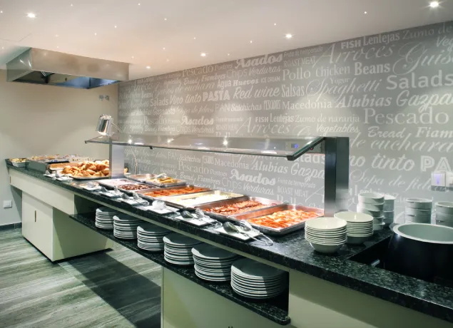 Buffet variado con platos calientes y fríos, estantes de platos, y fondo con letras decorativas.