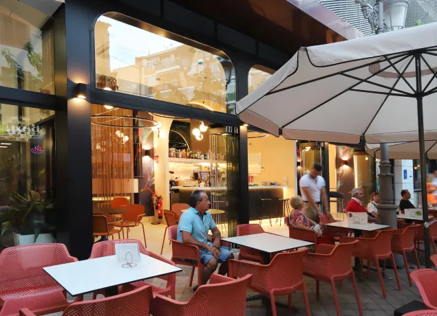 Terraza de café con mesas y sillas rojas, clientes sentados y gran sombrilla blanca.