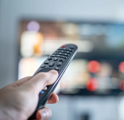 Mano sosteniendo un control remoto apuntando hacia un televisor desenfocado.