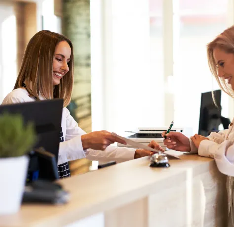 Recepcionista sonriente atendiendo a una mujer en un mostrador moderno.