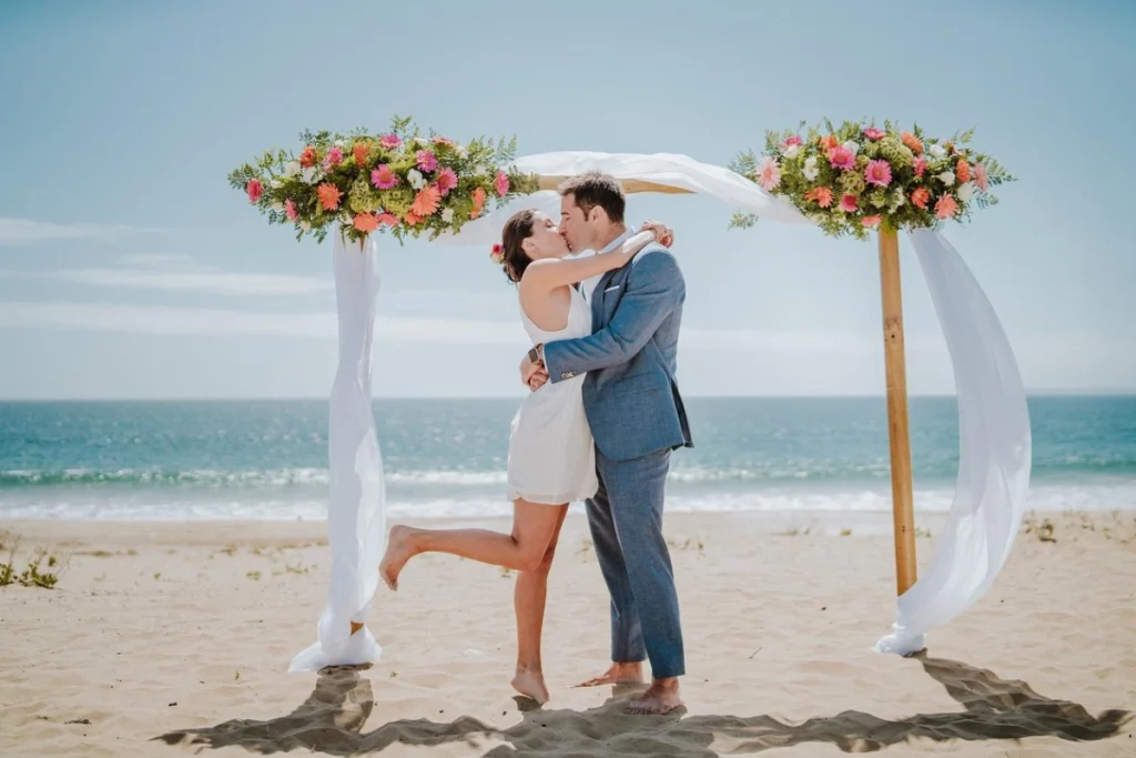 Couple kissing under floral arch on a sunny beach wedding ceremony.