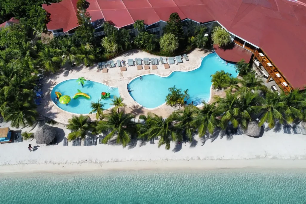 Piscinas y playa bordeadas de palmeras junto a un edificio con techo rojo.