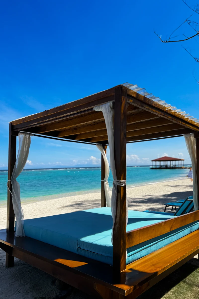 Cama de playa con dosel de madera frente al mar turquesa y cielo despejado.