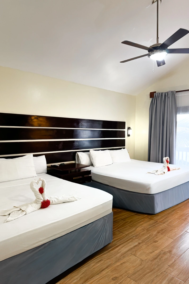 Habitación con dos camas, abanico en el techo y toallas dobladas.