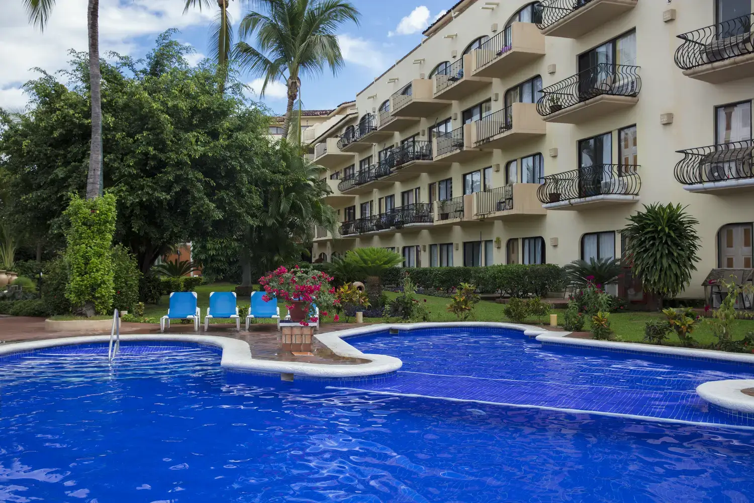 Piscina azul junto a un edificio de varios pisos con balcones, rodeada de plantas y palmeras. Hay tumbonas azules.
