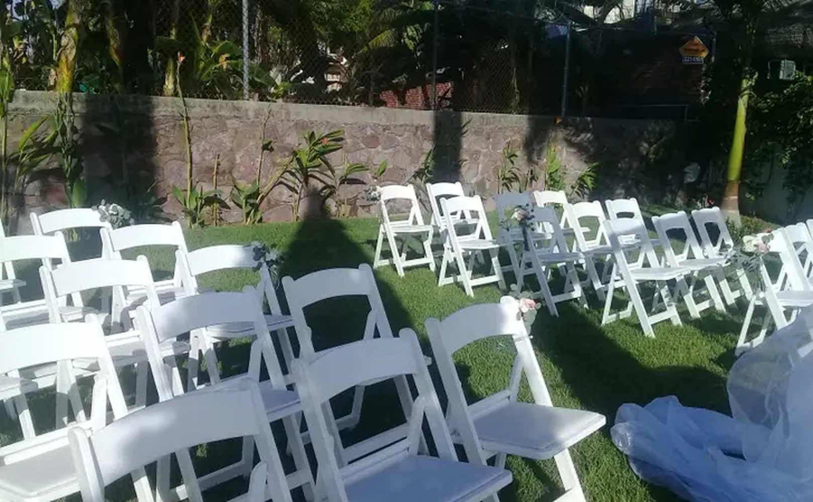 Sillas blancas dispuestas en un jardín al aire libre, preparadas para un evento, con plantas y sombra alrededor.