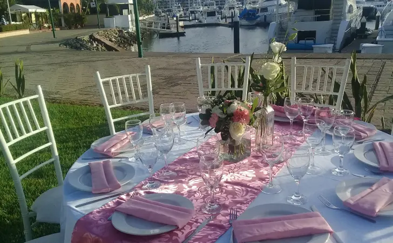 Mesa elegantemente decorada al aire libre, con manteles rosas, copas y flores, junto a un muelle con barcos.